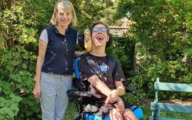 A smiling woman stands next to a young man in a motorized wheelchair with a therapy device on his head. They are outdoors in a garden with green plants, trees, and a green bench nearby.