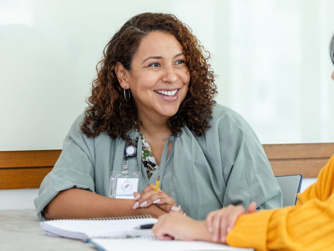 A woman in a green blouse with a lanyard smiles while talking to another woman in a yellow sweater. They sit at a table with notebooks, suggesting a friendly conversation or meeting.