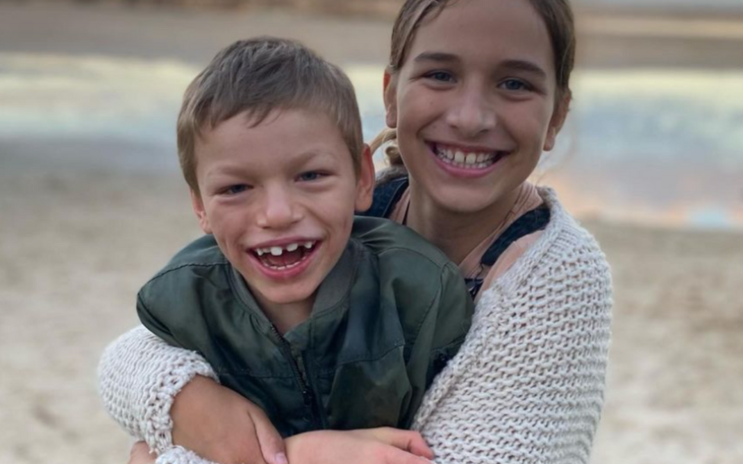 Two smiling children, a girl and a boy, hug each other on a sandy beach with water and a blurry horizon in the background. The boy wears a green jacket, and the girl wears a white knit sweater.