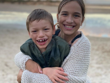 Two smiling children, a girl and a boy, hug each other on a sandy beach with water and a blurry horizon in the background. The boy wears a green jacket, and the girl wears a white knit sweater.