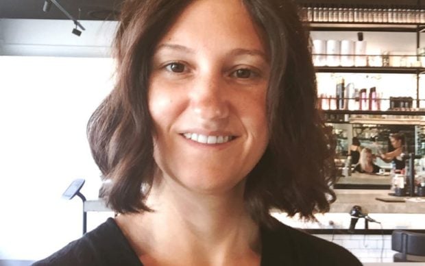 A woman with shoulder-length brown hair and a black top smiles at the camera in a modern, well-lit cafe with shelves and a mirrored wall in the background.