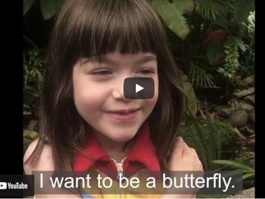 Young girl with brown hair smiles with her arms crossed over her chest, standing in front of green plants. Text at the bottom reads: I want to be a butterfly.