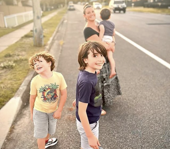 A woman stands on a street holding a toddler, while two young boys in casual clothes smile and look back at the camera. The sun sets in the background, casting a warm glow over the scene.