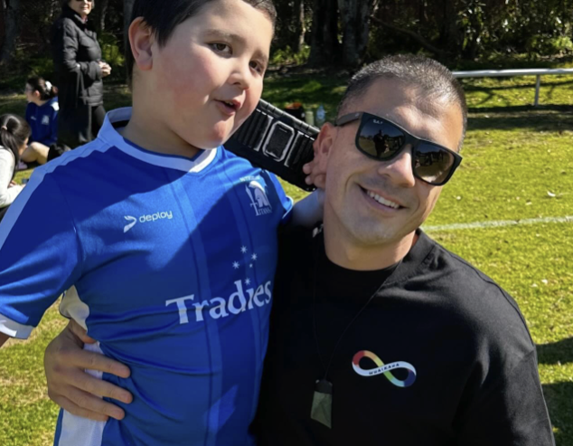 A smiling man wearing sunglasses and a black shirt with a rainbow infinity symbol holds a boy in a blue soccer jersey outdoors on a sunny day. Trees and people are visible in the background.