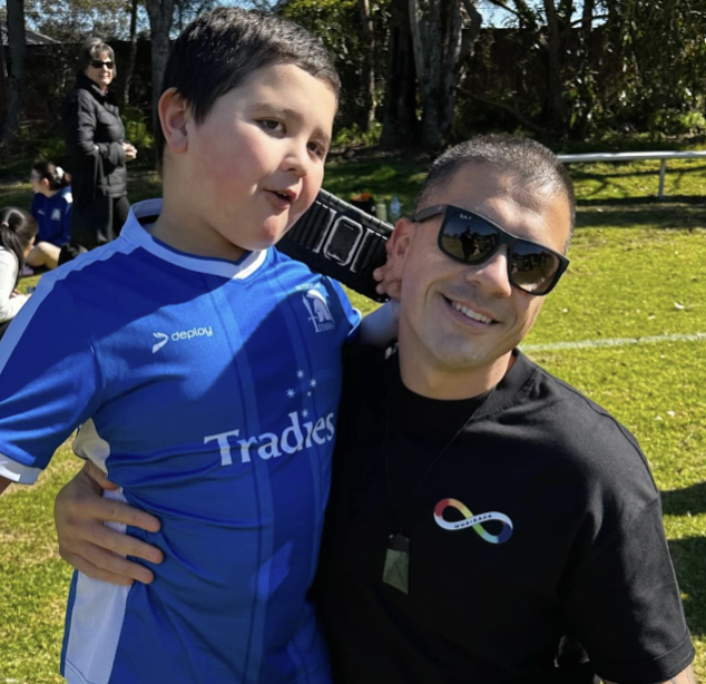 A smiling man wearing sunglasses and a black shirt with a rainbow infinity symbol holds a boy in a blue soccer jersey outdoors on a sunny day. Trees and people are visible in the background.