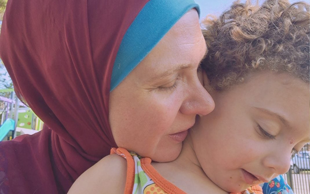 A woman in a maroon headscarf gently hugs a young child with curly hair, both enjoying a moment outdoors. The woman has her eyes closed and the child is looking down, wearing a colorful patterned shirt.