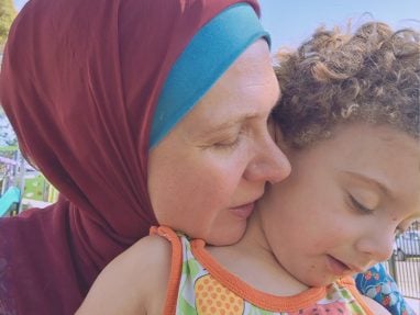 A woman in a maroon headscarf gently hugs a young child with curly hair, both enjoying a moment outdoors. The woman has her eyes closed and the child is looking down, wearing a colorful patterned shirt.