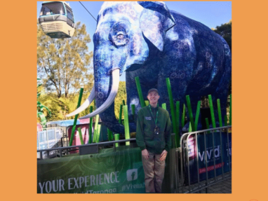 A person in a green jacket stands smiling in front of a large blue elephant sculpture at an outdoor event. A cable car and trees are visible in the background. The image has an orange border with “the good life” written at the bottom.