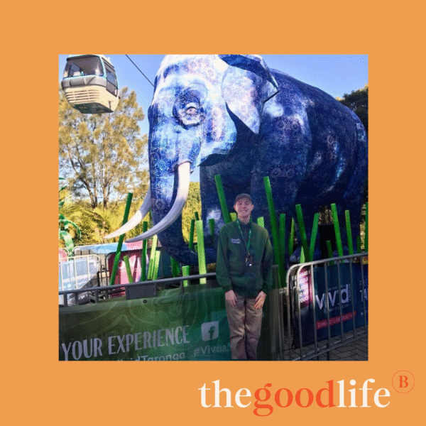 A person in a green jacket stands smiling in front of a large blue elephant sculpture at an outdoor event. A cable car and trees are visible in the background. The image has an orange border with “the good life” written at the bottom.