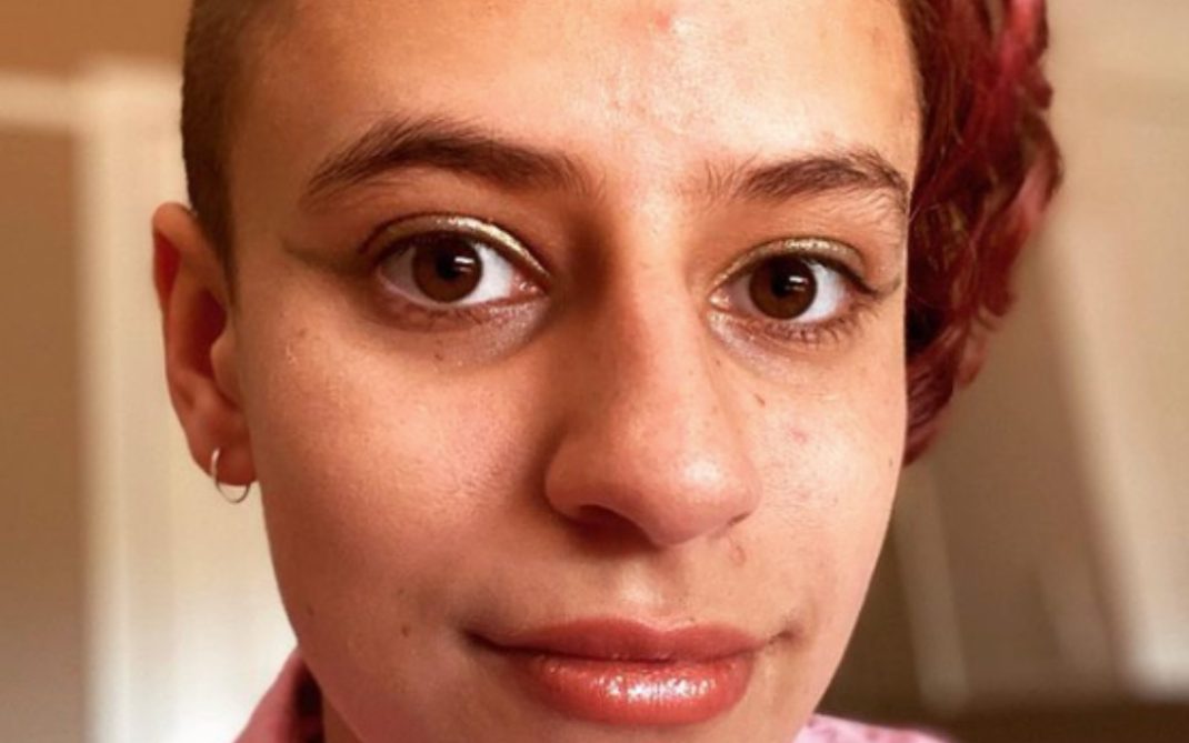 A person with short, partially shaved hair and a hint of red coloring on one side, wearing a pink collared shirt, looks directly at the camera with a neutral expression.