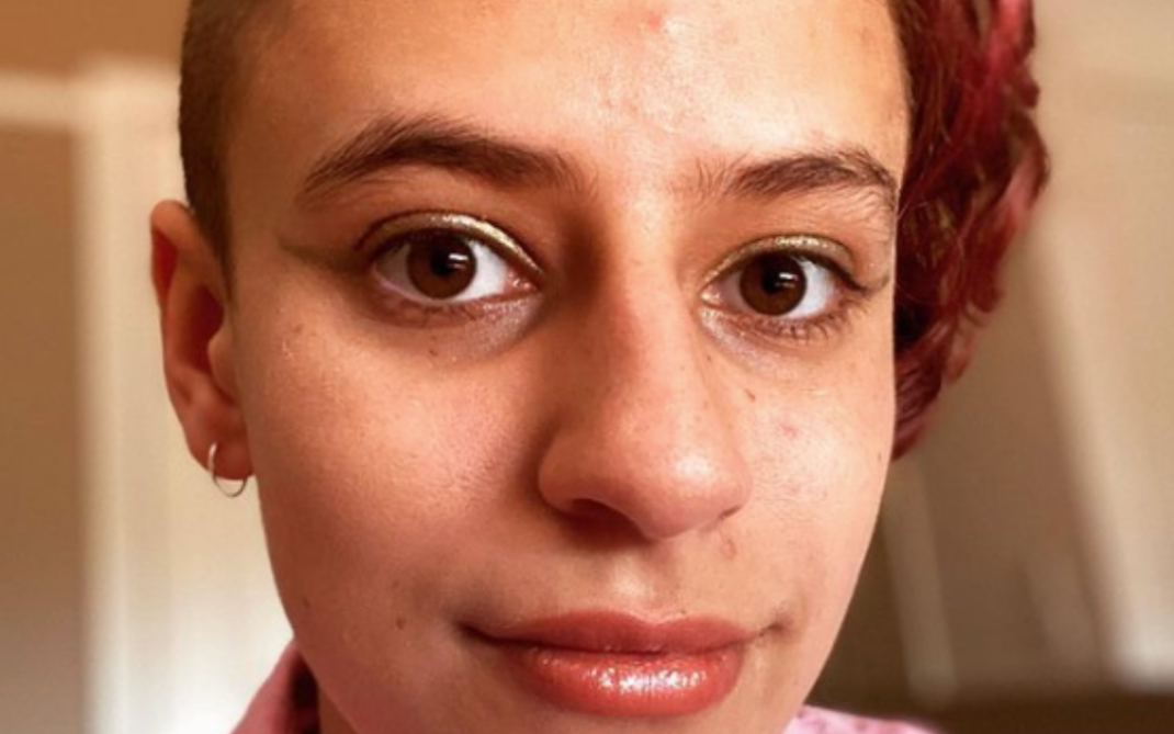 A person with short, partially shaved hair and a hint of red coloring on one side, wearing a pink collared shirt, looks directly at the camera with a neutral expression.