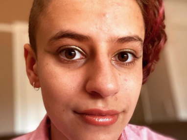 A person with short, partially shaved hair and a hint of red coloring on one side, wearing a pink collared shirt, looks directly at the camera with a neutral expression.