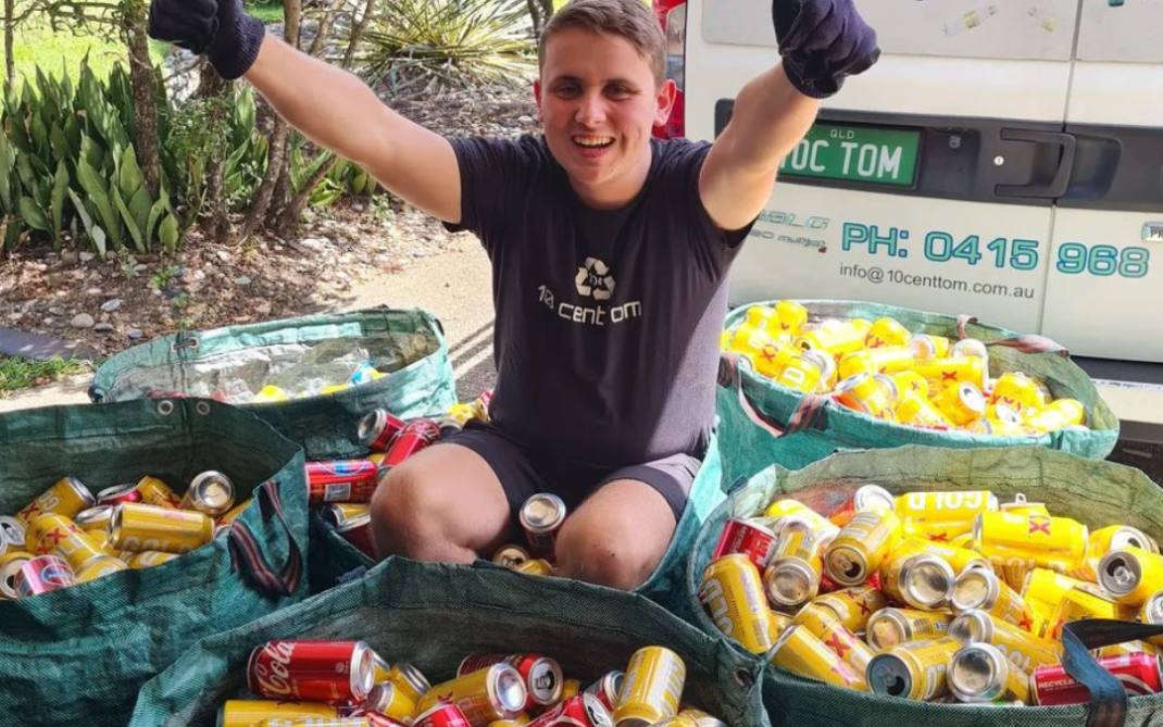 A smiling person in black gloves and a dark shirt sits among large bags full of empty drink cans, giving two thumbs up. A van with recycling signs and contact info is parked behind them.