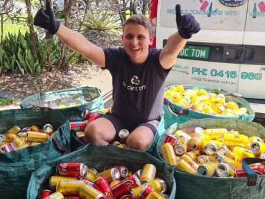 A smiling person in black gloves and a dark shirt sits among large bags full of empty drink cans, giving two thumbs up. A van with recycling signs and contact info is parked behind them.