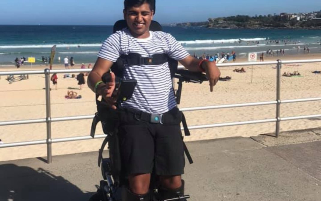 A man in a striped shirt uses a standing wheelchair on a beachside promenade, smiling at the camera. The ocean, sandy beach, and people relaxing are visible in the background under a clear blue sky.