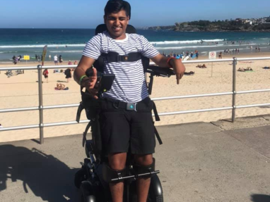 A man in a striped shirt uses a standing wheelchair on a beachside promenade, smiling at the camera. The ocean, sandy beach, and people relaxing are visible in the background under a clear blue sky.