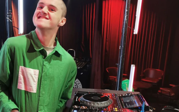 A smiling person in a green shirt stands beside DJ equipment, with colorful lights and empty chairs visible in the background.