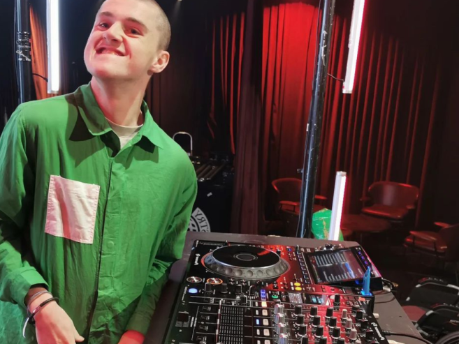A smiling person in a green shirt stands beside DJ equipment, with colorful lights and empty chairs visible in the background.