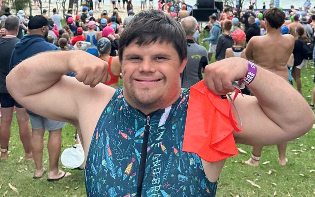 A young man in a colorful wetsuit smiles and flexes his arms, holding an orange swim cap. He stands on grass near a crowd gathered by a lakeshore, with swimmers and kayaks visible in the water.