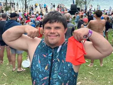 A young man in a colorful wetsuit smiles and flexes his arms, holding an orange swim cap. He stands on grass near a crowd gathered by a lakeshore, with swimmers and kayaks visible in the water.