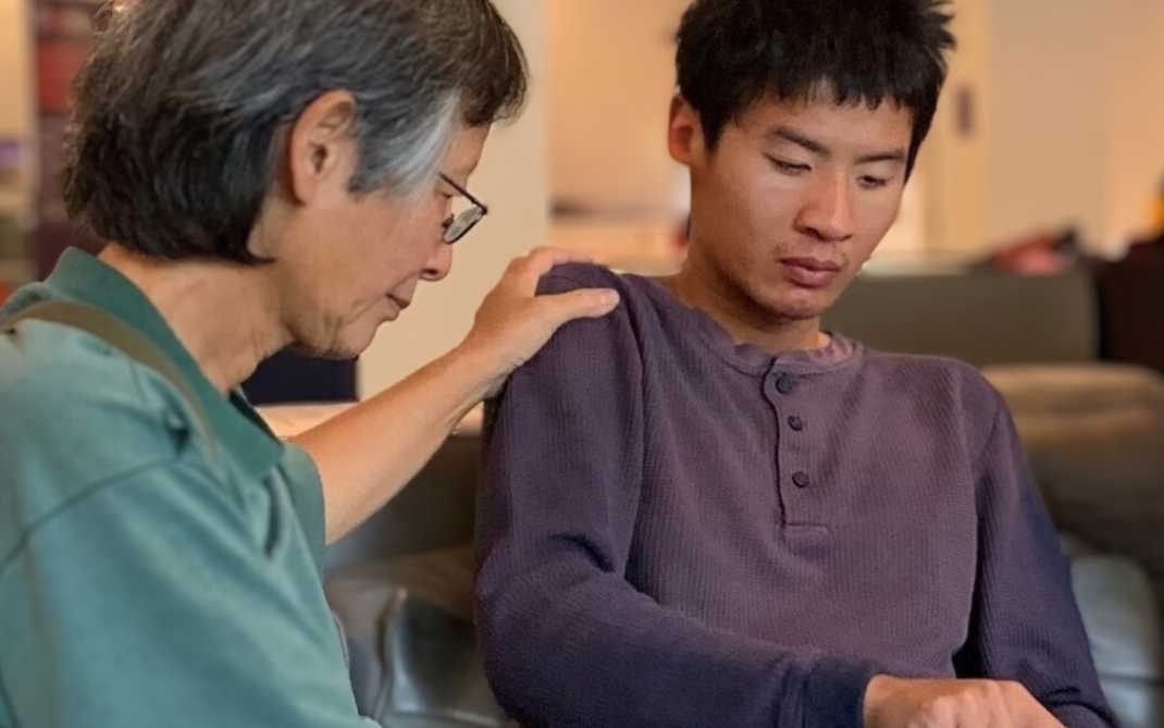An older person gently rests their hand on the shoulder of a young man, who is sitting on a couch and using a tablet. Both appear focused, sharing a quiet moment in a well-lit indoor setting.