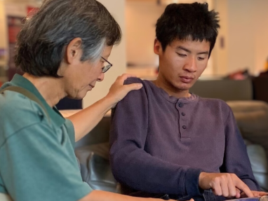 An older person gently rests their hand on the shoulder of a young man, who is sitting on a couch and using a tablet. Both appear focused, sharing a quiet moment in a well-lit indoor setting.
