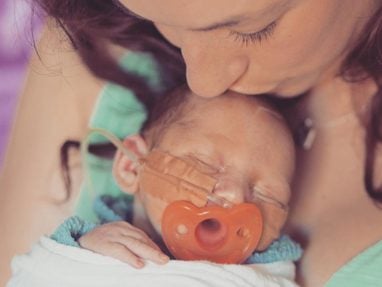 A woman gently kisses the forehead of a sleeping newborn who has medical tape and a tube on their face, and is holding an orange pacifier. The baby is wrapped in a blanket, showing a moment of tenderness and care.
