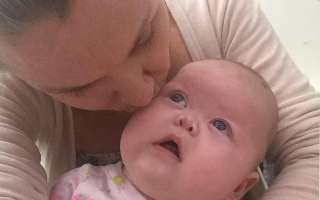 A woman kisses the forehead of a baby who is looking up at her with wide eyes. Both are dressed in light-colored clothing. The image captures a tender, affectionate moment.