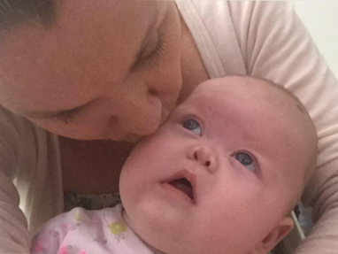 A woman kisses the forehead of a baby who is looking up at her with wide eyes. Both are dressed in light-colored clothing. The image captures a tender, affectionate moment.