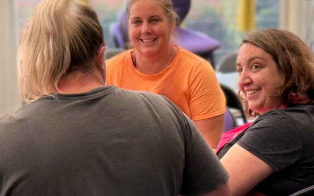 Three women sit together indoors, smiling and chatting. One woman in an orange shirt faces the camera, while the other two, wearing dark shirts, are turned slightly away, engaged in conversation. Bright windows and greenery are visible behind them.