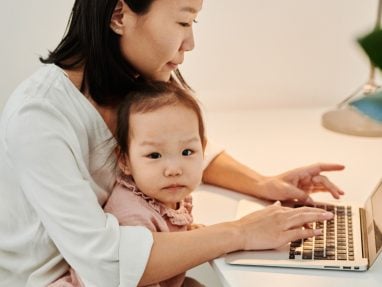A woman sits at a desk working on a laptop with a young child in her lap. The child looks at the camera while the woman focuses on the computer. The setting appears to be a home office.