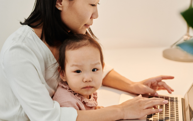 A woman sits at a desk working on a laptop with a young child in her lap. The child looks at the camera while the woman focuses on the computer. The setting appears to be a home office.