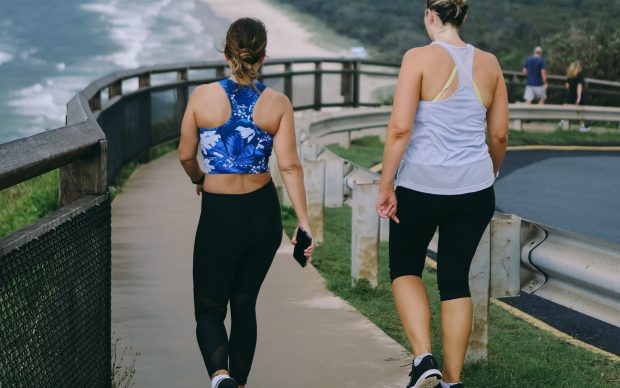 Two women in athletic wear walk on a paved path next to a beach, with ocean waves and greenery visible in the background. One holds a phone, and both appear to be exercising or enjoying a casual walk.