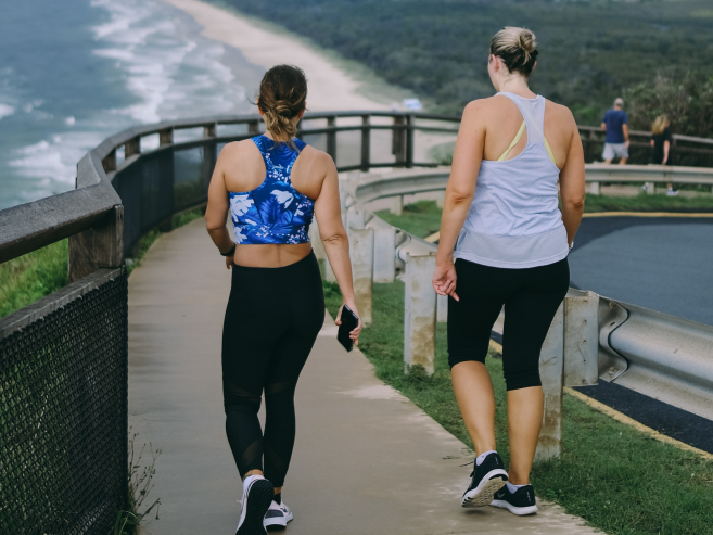 Two women in athletic wear walk on a paved path next to a beach, with ocean waves and greenery visible in the background. One holds a phone, and both appear to be exercising or enjoying a casual walk.