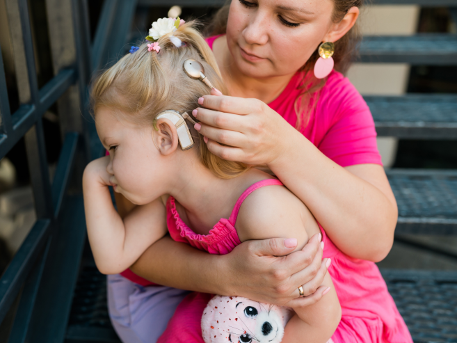 A woman gently adjusts a cochlear implant on a young girl’s head as they sit on outdoor steps. The girl, wearing a pink dress and holding a stuffed toy, looks down while the woman embraces her.