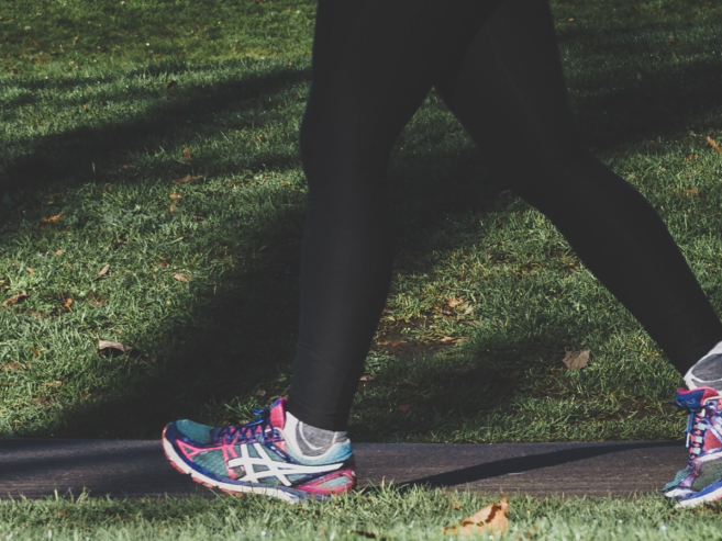 A person wearing black leggings and colorful running shoes walks on a paved path next to a grassy area with shadows from trees.