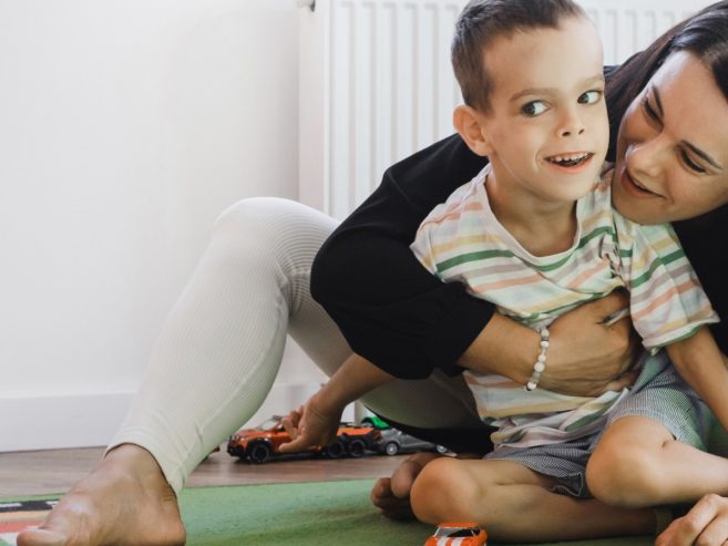 A woman lovingly hugs a smiling young boy while sitting on a green mat in a playroom, surrounded by toy cars and trucks.