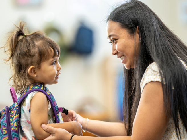 A woman smiles warmly at a young girl wearing a backpack. The woman is holding the girls arms gently, and they appear to be having a caring conversation in a bright, indoor setting.