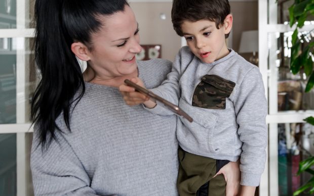 A woman holds a young boy while they both look at a tablet. The woman is smiling, and the boy appears focused on the screen. They are indoors in a cozy, well-lit room.