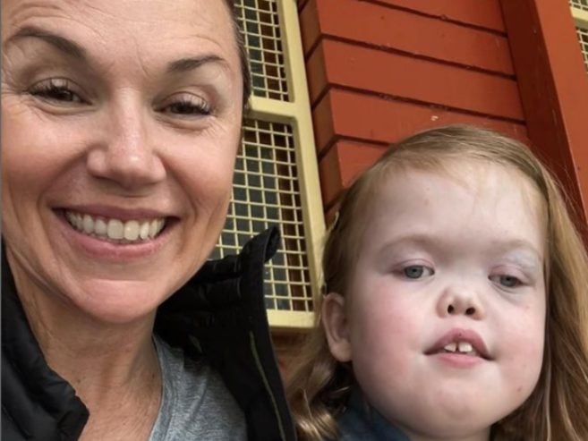 A woman smiling next to a young girl with long light brown hair, in front of a red and yellow building with gridded windows.