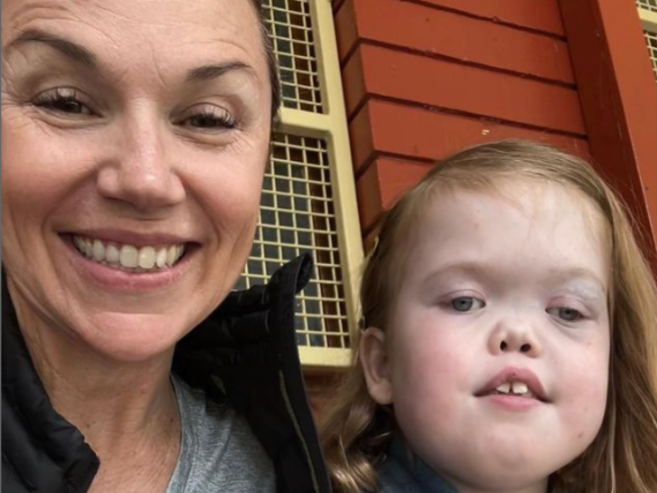 A woman smiling next to a young girl with long light brown hair, in front of a red and yellow building with gridded windows.