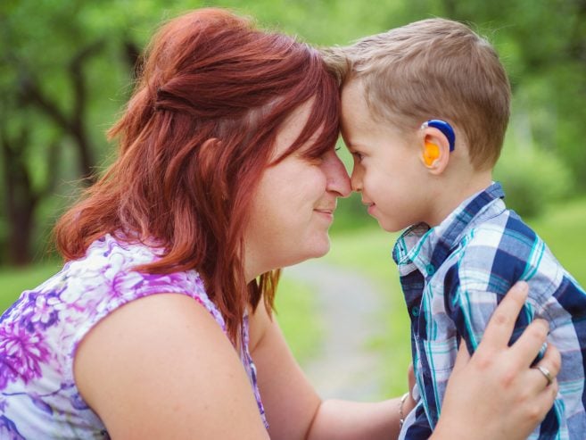 A woman and a young boy with a hearing aid touch foreheads and smile at each other outdoors, surrounded by greenery. The moment appears warm and affectionate.