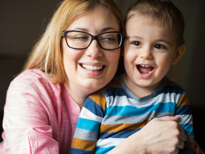 A smiling woman with glasses hugs a happy young boy in a striped shirt. They are sitting close together, both looking slightly off-camera, enjoying a joyful moment indoors.