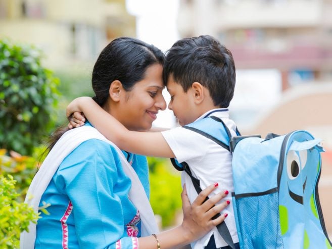 A mother and her young son, wearing a blue school uniform and backpack, share a warm embrace and smile outdoors before school, their foreheads touching affectionately.