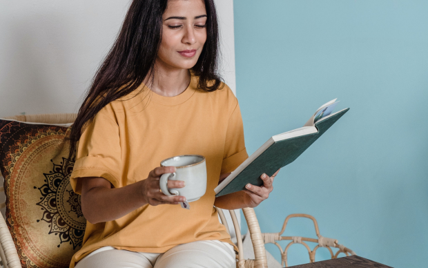 A woman with long dark hair sits in a chair, holding a mug in one hand and reading a book with the other. She wears a yellow shirt and light pants, with a decorative pillow behind her and a wooden table in front.