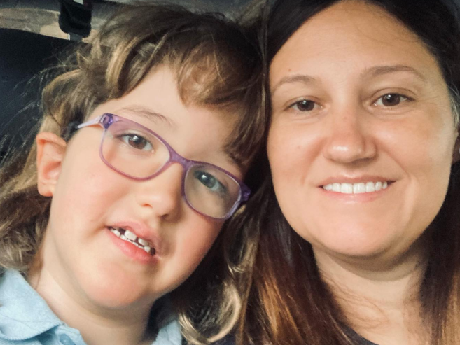 A young girl with glasses and a woman with long brown hair are smiling together, their heads close, in a casual selfie.