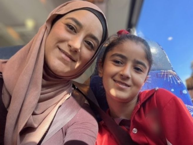 A woman in a light brown hijab and a young girl in a red shirt smile while sitting together on a bus, with blue sky visible through the window behind them.