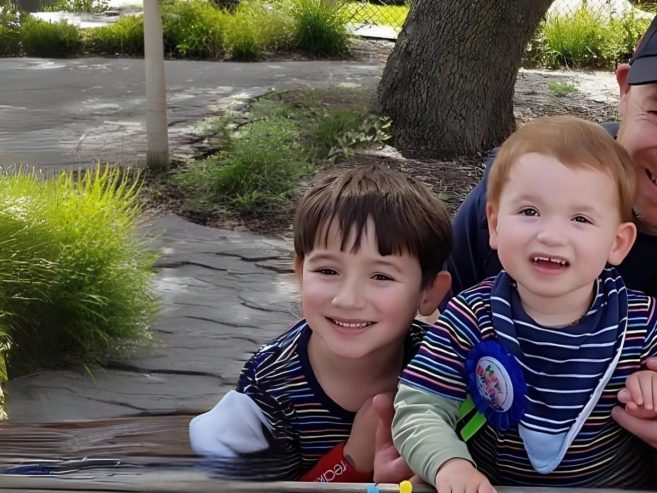 A family of four, including two young children and two adults, sit closely together outdoors, smiling and laughing near greenery and a large tree.