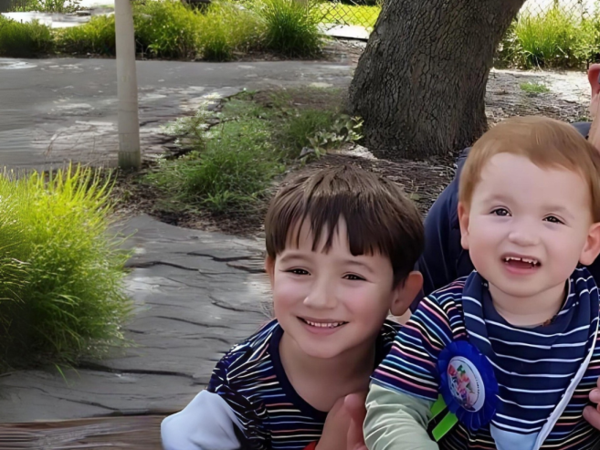 A family of four, including two young children and two adults, sit closely together outdoors, smiling and laughing near greenery and a large tree.