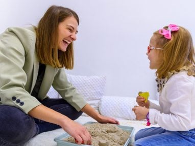 An adult woman and a young girl sit on the floor, smiling and playing with kinetic sand in a tray. The girl wears glasses and a pink bow, and both appear happy and engaged in the activity.